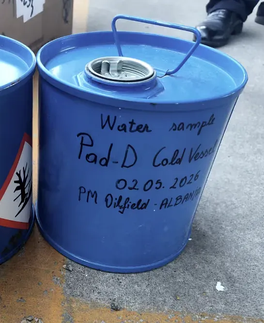 A steel drum opened for sampling, hand-marked with a sample identification, a corrosive hazard diamond on the drum beside it